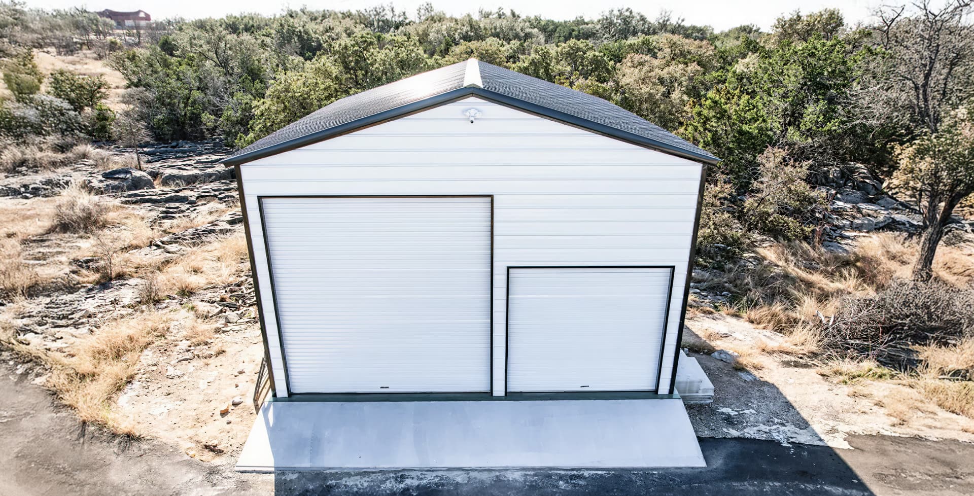 A tall white metal garage with black trim and two roll-up doors, one extra-high for an RV, located on a raised concrete foundation.