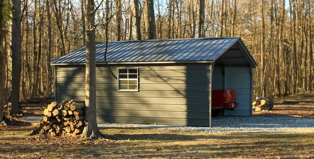 A gray metal garage with a red truck parked inside, surrounded by trees and stacks of firewood.