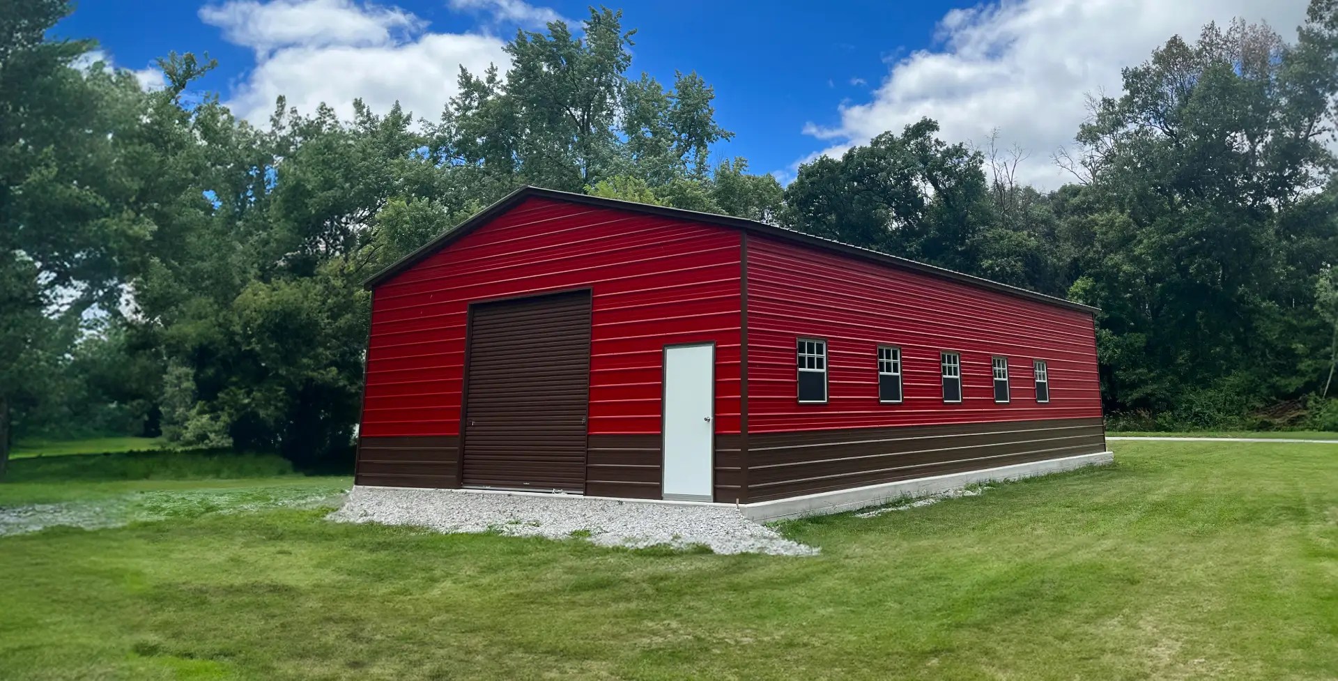A large red and brown metal garage with multiple windows, a white door, and a brown roll-up door, set on a grassy lot with trees in the background.