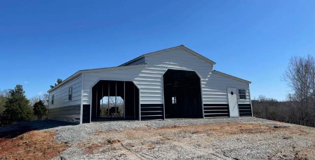A white metal barn with black lower trim sits atop a gravel-covered hill. The structure has large open bays in front, visible framing inside, and a white entry door with a small window on the right side.