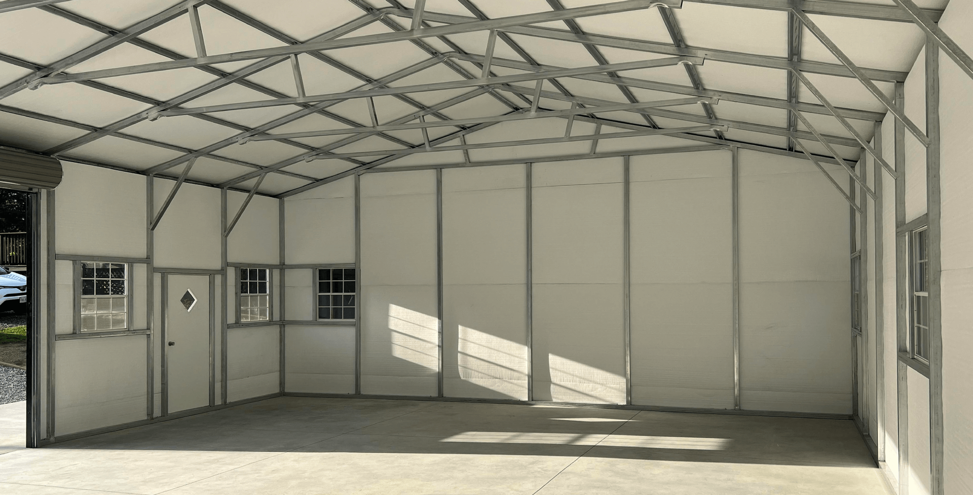 Interior view of a metal-framed barndominium shell. The structure has exposed steel trusses across the ceiling, steel wall framing, cream-colored wall panels, and several rectangular windows.
