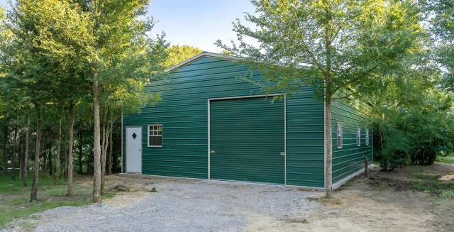 A large green metal garage building set among tall trees, with a gravel driveway leading to a white entry door and a closed roll-up garage door on the front.
