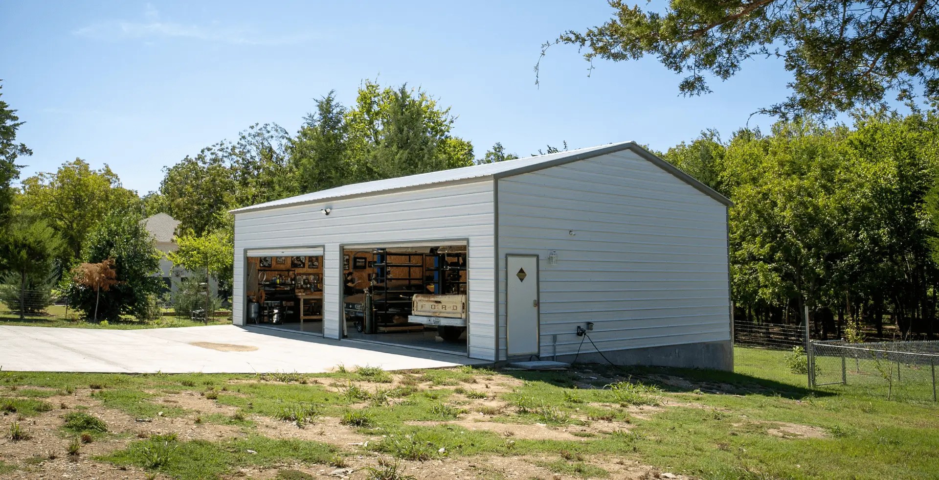 A white metal garage building with two open roll-up bays revealing workshop tools and a vintage Ford truck inside. The structure sits beside a concrete driveway and is surrounded by grass, trees, and a wire fence.
