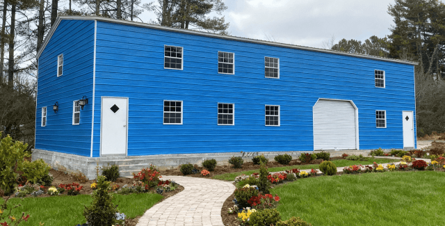 Exterior view of a bright blue two-story barndominium with multiple evenly spaced white-trimmed windows. It features two white entry doors—one on each end—and a large white garage-style door. A landscaped garden with a curved stone pathway leads toward the building.