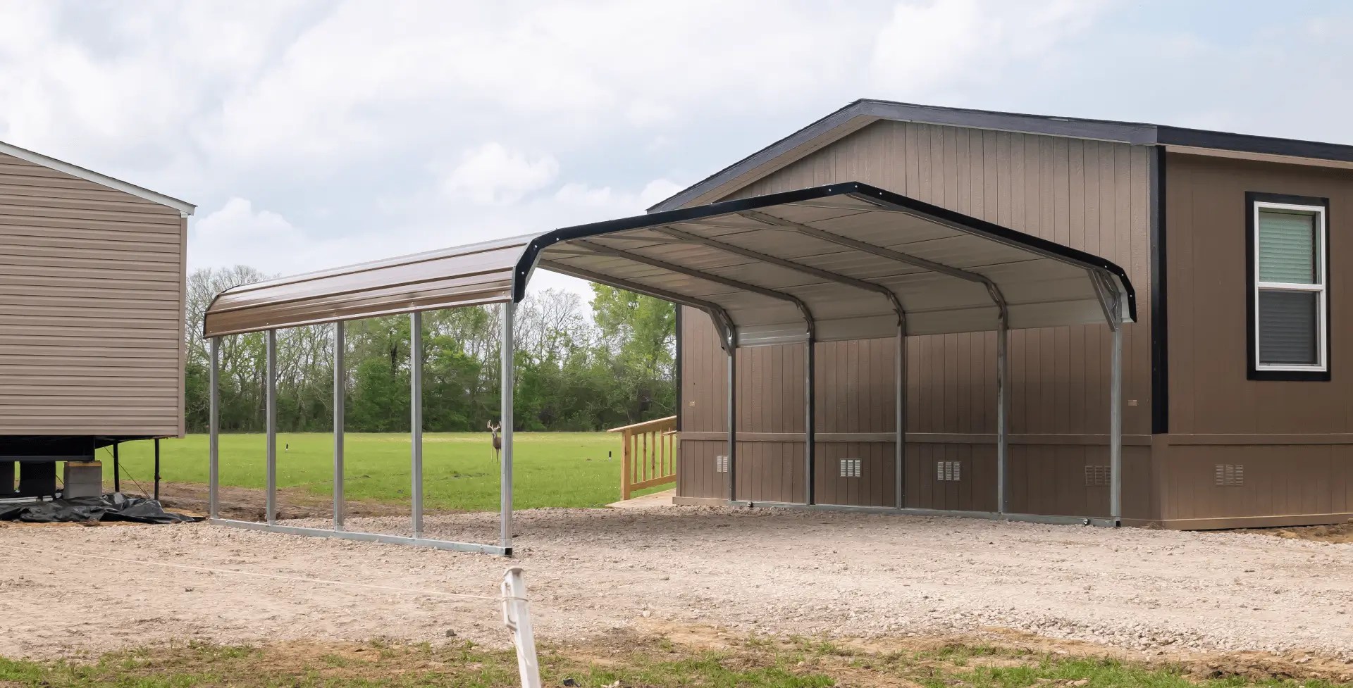Large metal carport installed beside a brown manufactured home. The structure has a tall, peaked metal roof supported by steel posts, with open sides and a gravel base. A second smaller carport stands nearby on a dirt pad.