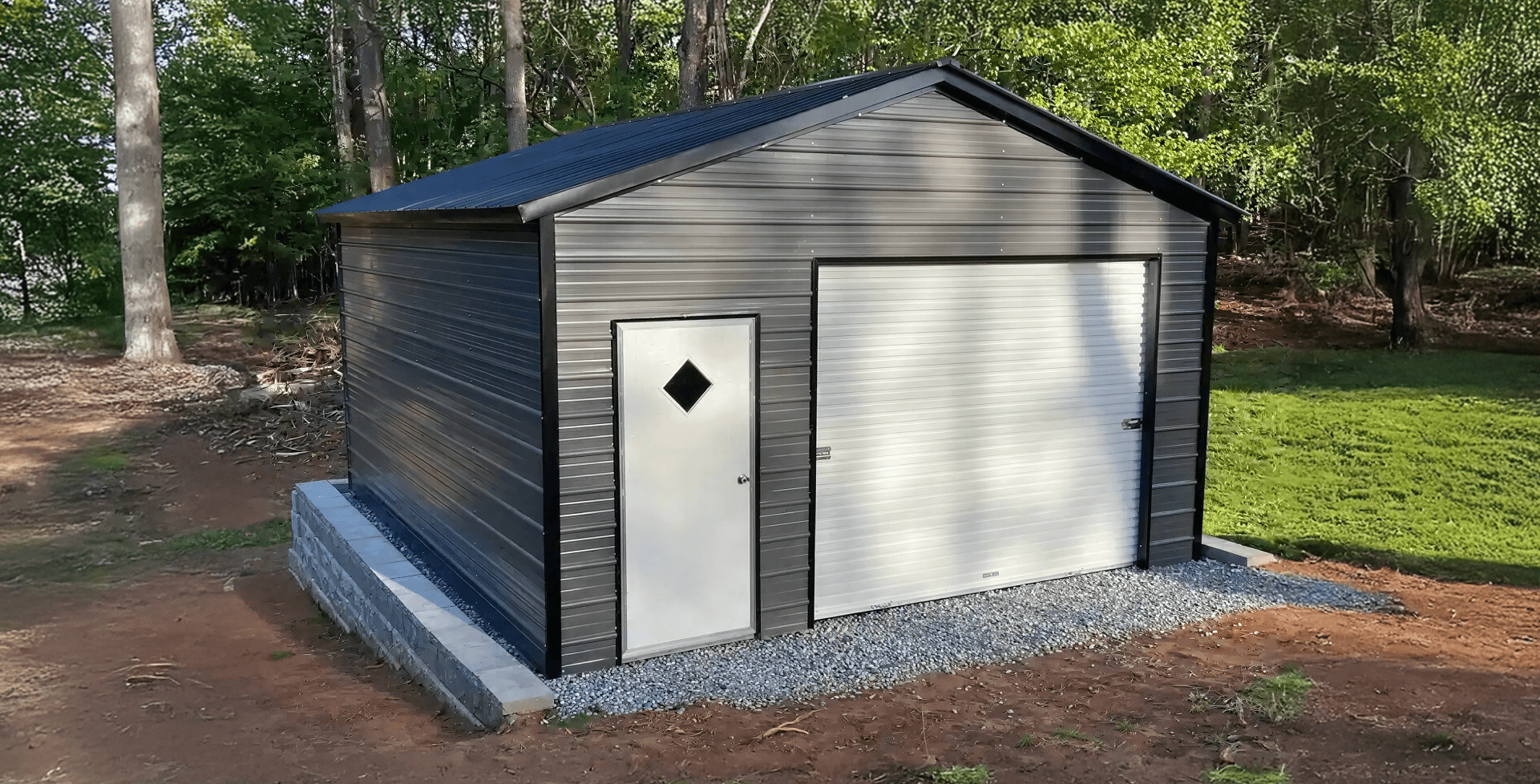 Small black metal garage with a gabled roof, a white side-entry door with a diamond window, and a single roll-up garage door. The building sits on a raised concrete block foundation with gravel in front.
