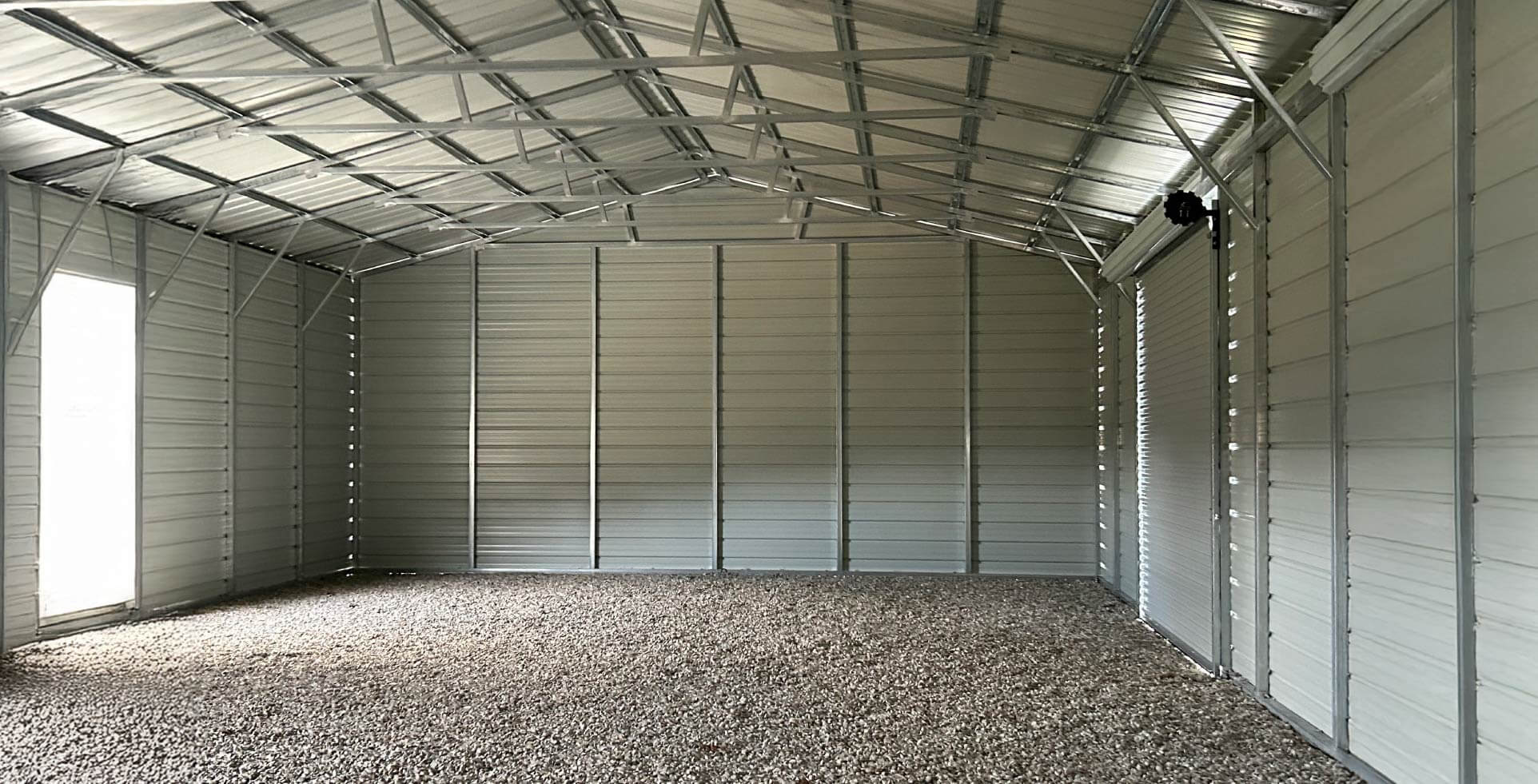 The inside of a large metal barn with gravel flooring and steel framing. Light enters from a door opening on the left and through small gaps along the metal wall panels, illuminating the open interior and high gabled roof structure.