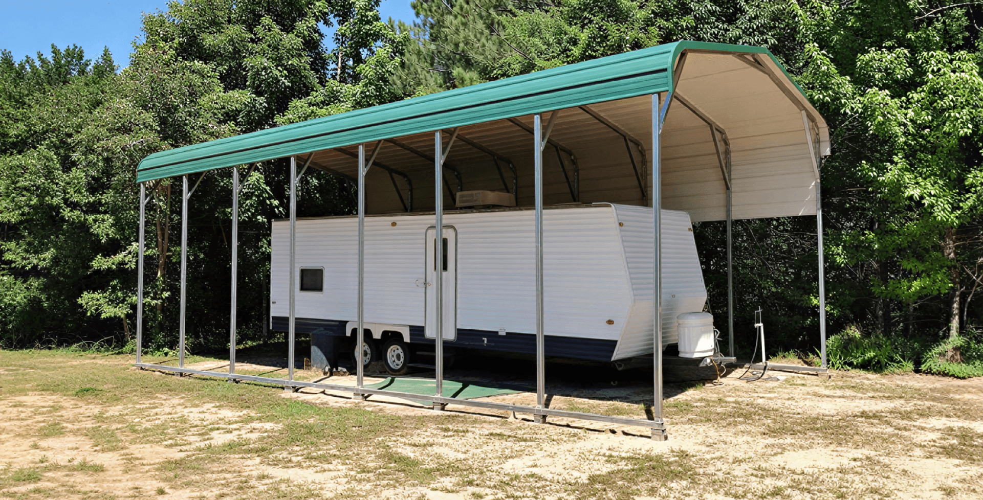 Tall metal RV carport with a green rounded roof sheltering a long white travel trailer. The structure has open sides supported by slim steel posts and is set in a clearing surrounded by dense trees on a slightly sandy patch of ground.