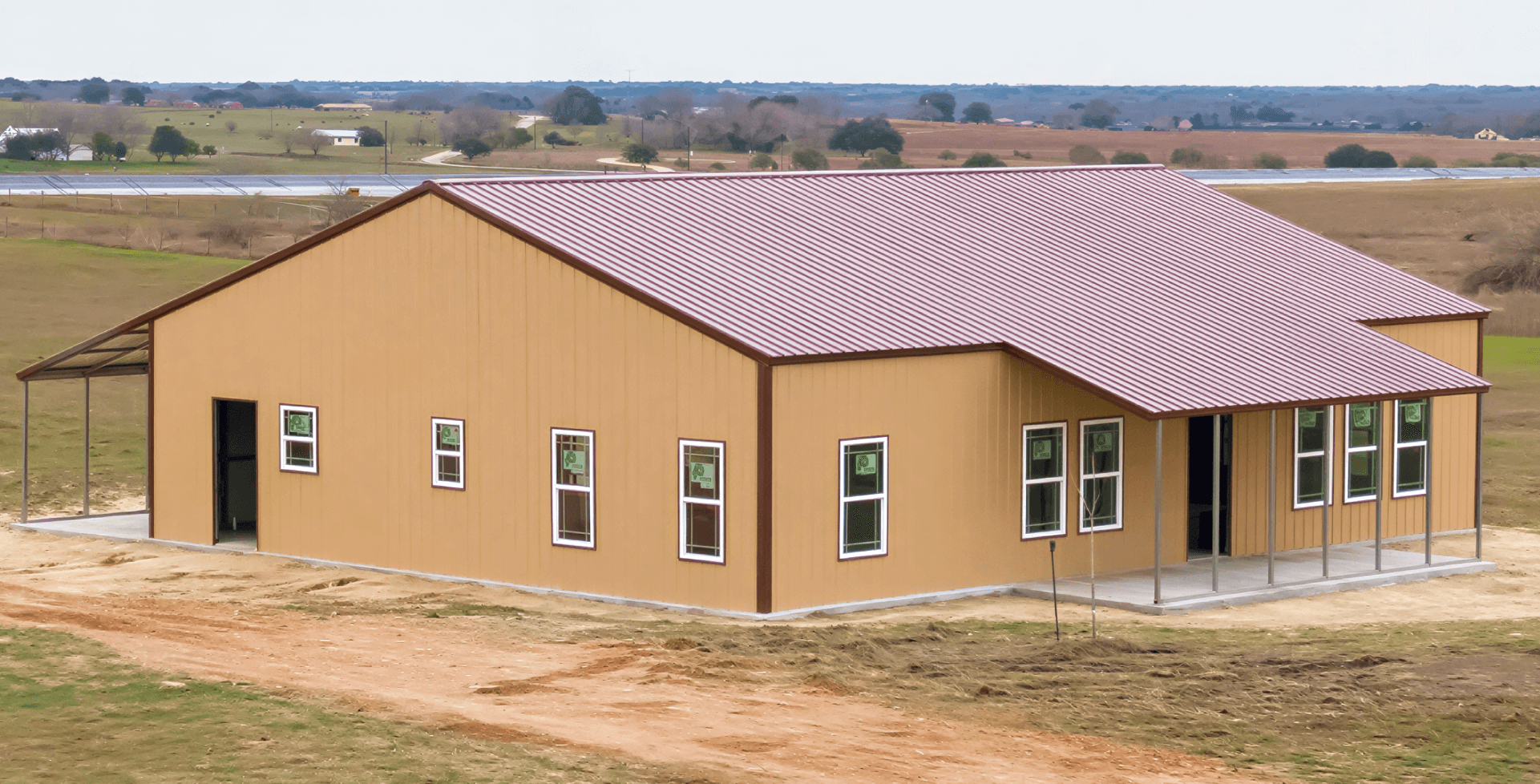 Large tan metal building, featuring many windows and a burgundy metal roof. The structure stands on a concrete slab.