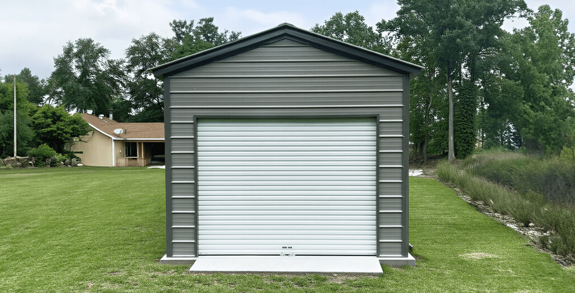 Gray metal garage viewed straight-on, centered in the frame. It features a single white roll-up door and sits on a concrete pad in a well-maintained grassy yard.