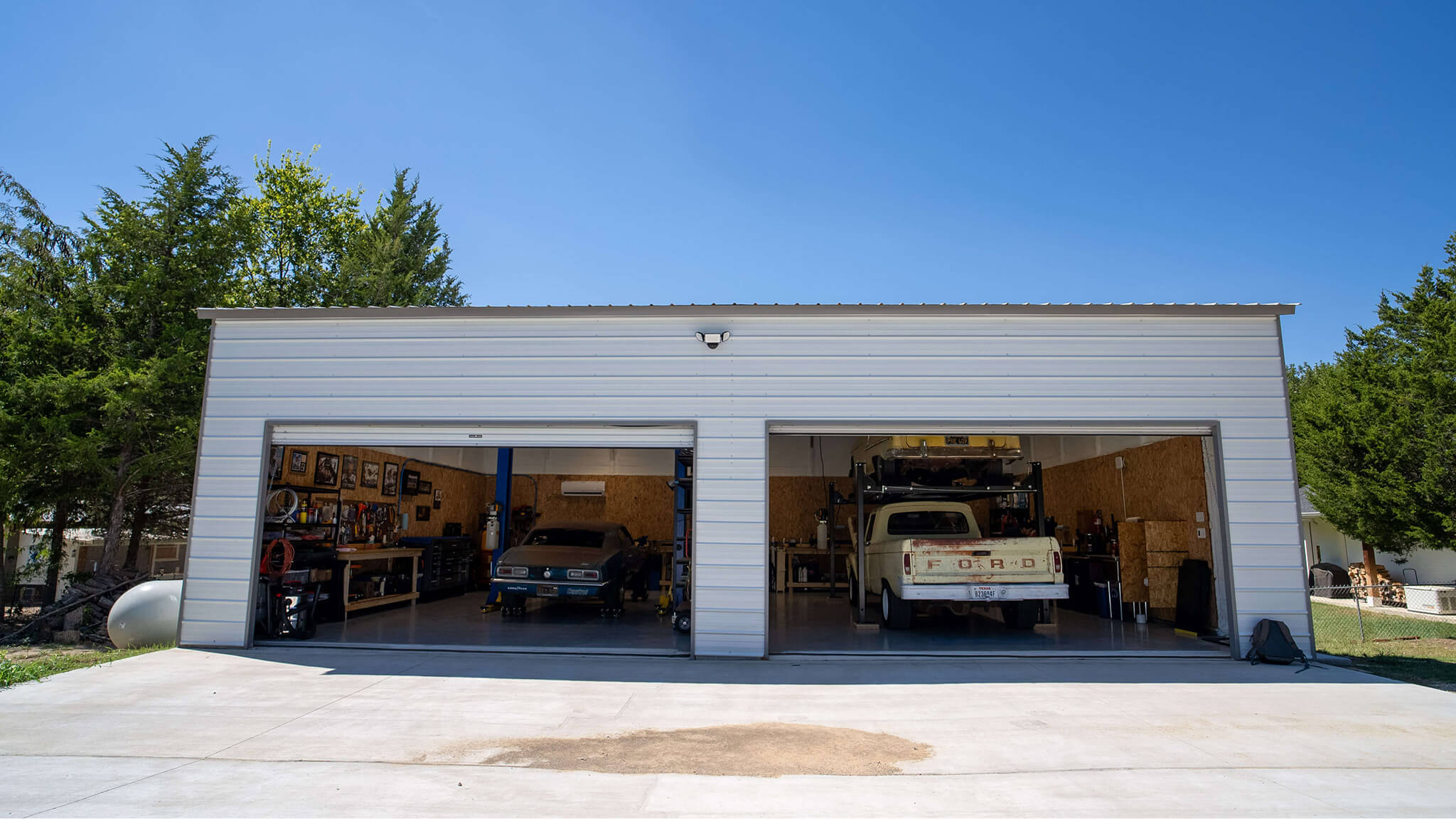 White 30x40 metal garage with two large open bay doors, revealing a fully equipped mechanic workspace inside with vehicles, tool benches, shelving, and lifts, set on a concrete driveway.