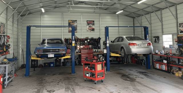 A wide interior view of a mechanic workshop inside a metal building. Two cars—one blue classic car and one silver sedan—are elevated on separate vehicle lifts. Workbenches, tool storage, posters, and various automotive equipment line the walls under a high, open steel truss ceiling.