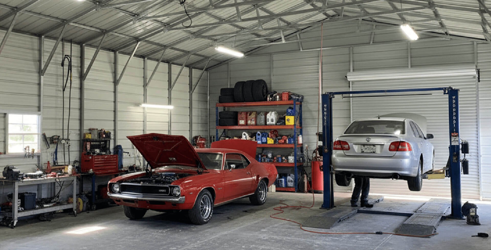Inside a mechanic shop, a red classic muscle car with its hood open sits beside a silver sedan lifted on a hydraulic car lift. Shelves filled with tools, tires, and containers line the back wall, all inside a large metal building with exposed steel framing and bright overhead lighting.
