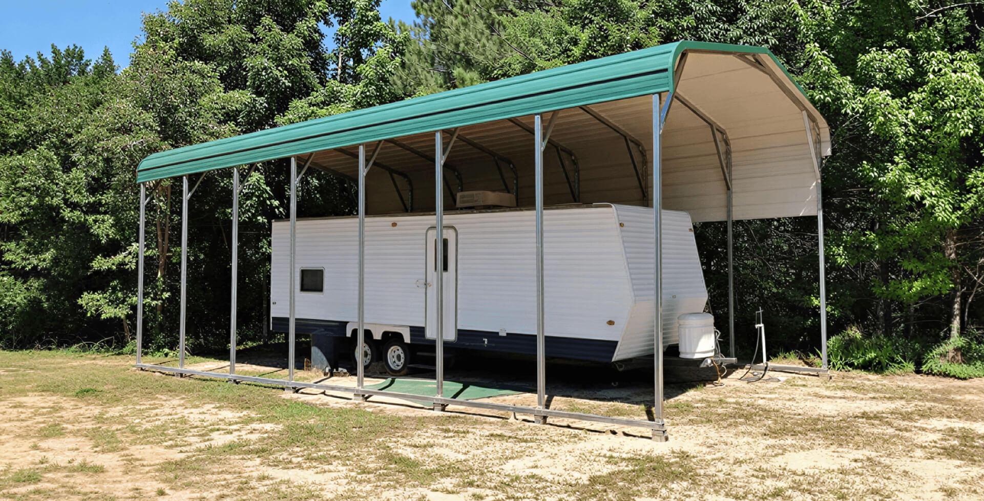 Tall metal RV carport with a green rounded roof sheltering a long white travel trailer. The structure has open sides supported by slim steel posts and is set in a clearing surrounded by dense trees on a slightly sandy patch of ground.