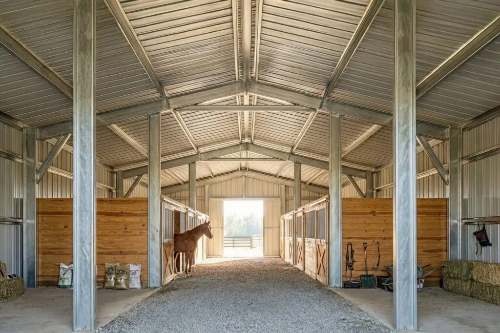 Interior of a metal horse barn with steel beams, gravel aisle, and wooden horse stalls, showing a horse standing in a stall with natural light entering from the open end.