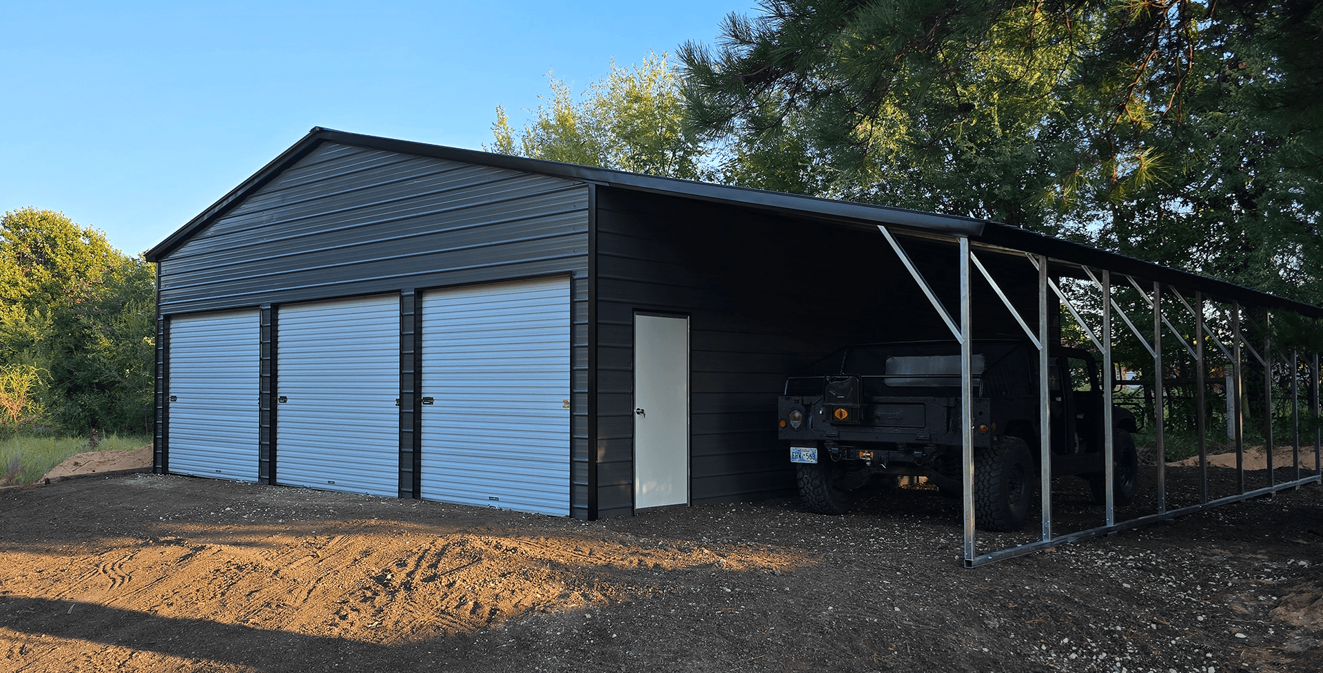 Metal garage with three white roll-up doors and a side entry door, attached to an open carport sheltering a black off-road vehicle. The structure sits on a gravel lot surrounded by trees under a clear blue sky.