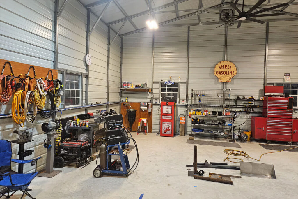 Spacious metal workshop interior with concrete floor, workbenches along the walls, red tool chest, air compressor, welding machine, and coiled extension cords. A vintage “Shell Gasoline” sign hangs on the wall above shelves of tools and supplies, with a large ceiling fan and bright overhead lights.