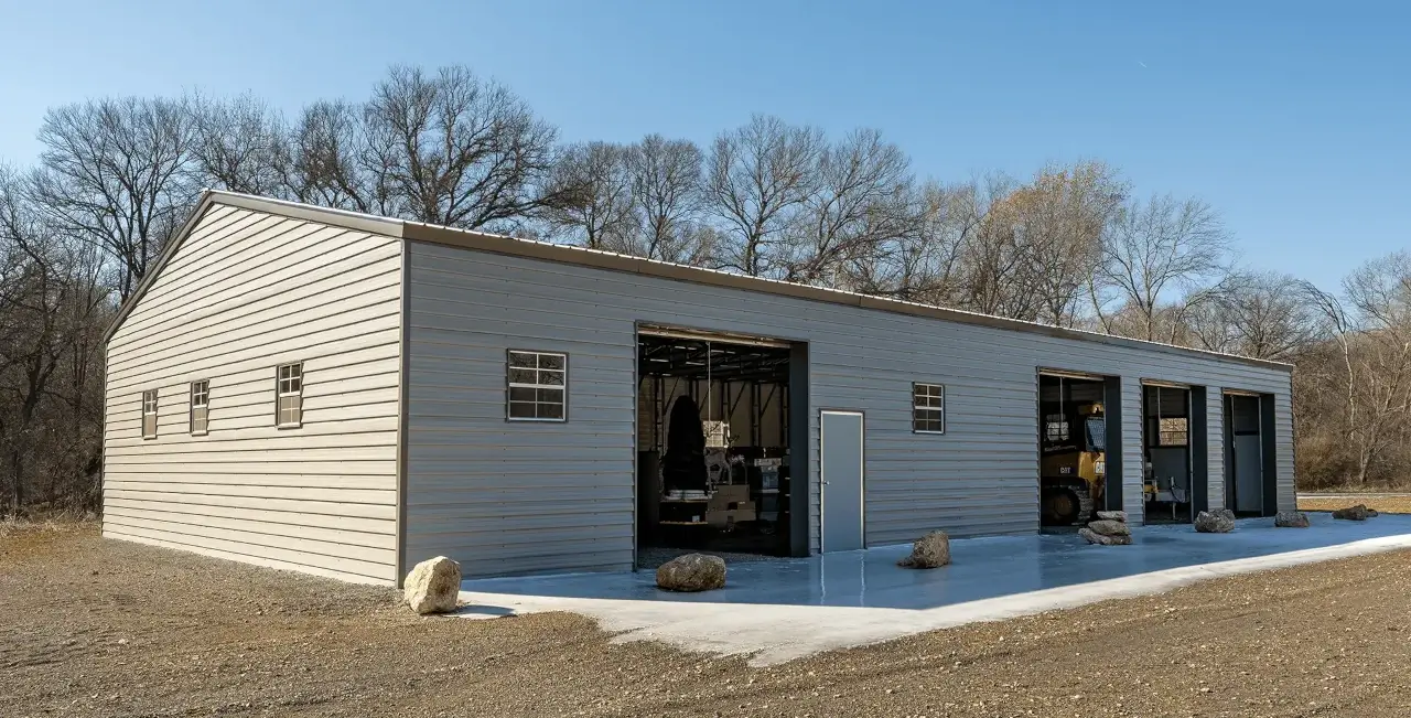 Metal-sided rectangular workshop building with a sloped roof and three open garage bays on a concrete slab. Construction equipment and storage items are visible inside, with leafless trees and a clear blue sky in the background.