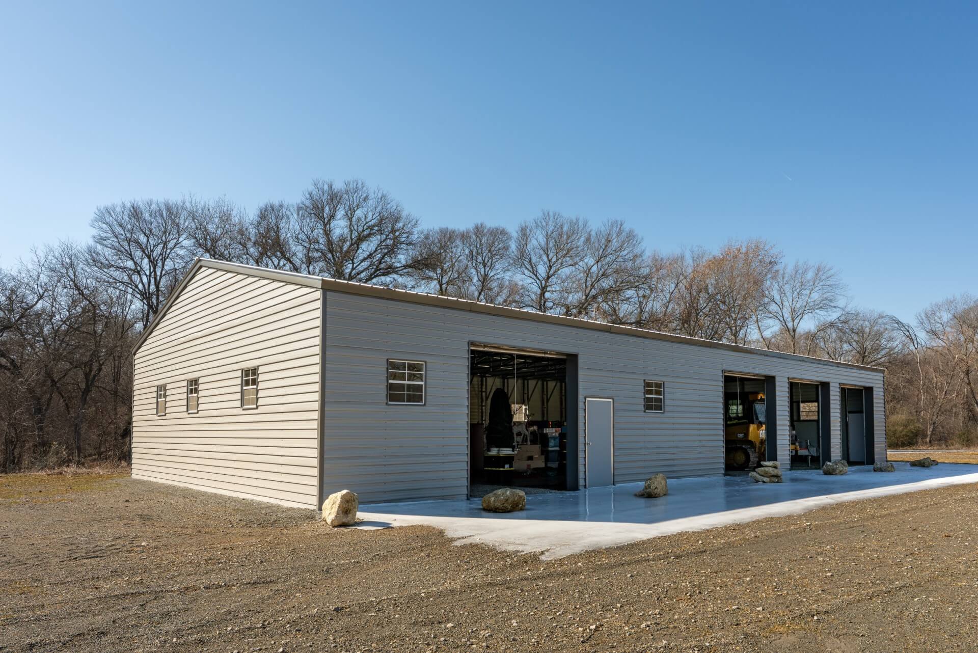 Large beige metal garage building with three open bay doors and a side entry door on a concrete slab, set on a gravel lot with leafless trees in the background. Equipment and storage items are visible inside the open bays under a clear blue sky.
