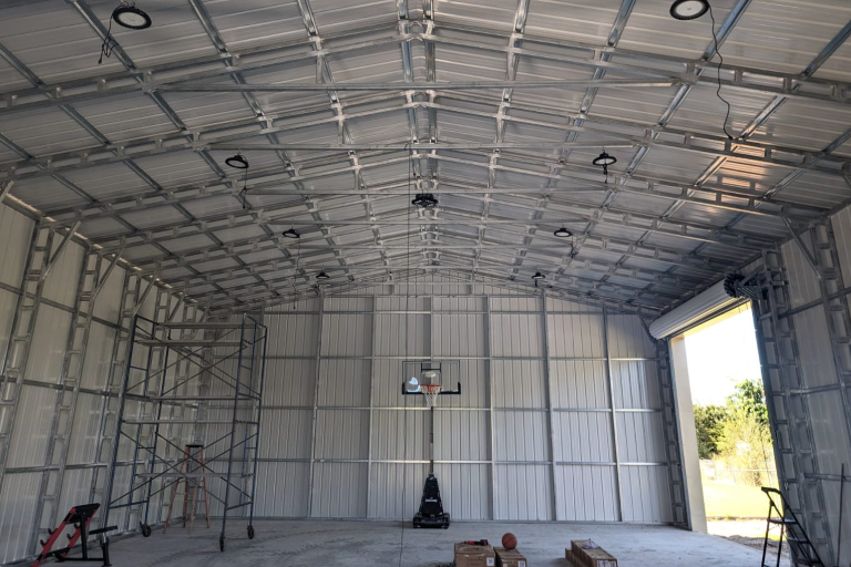 Interior of a large metal building with exposed steel framing and corrugated wall panels, viewed from the open garage door. A basketball hoop is mounted on the far wall above a concrete floor, with scaffolding and a few tools inside.