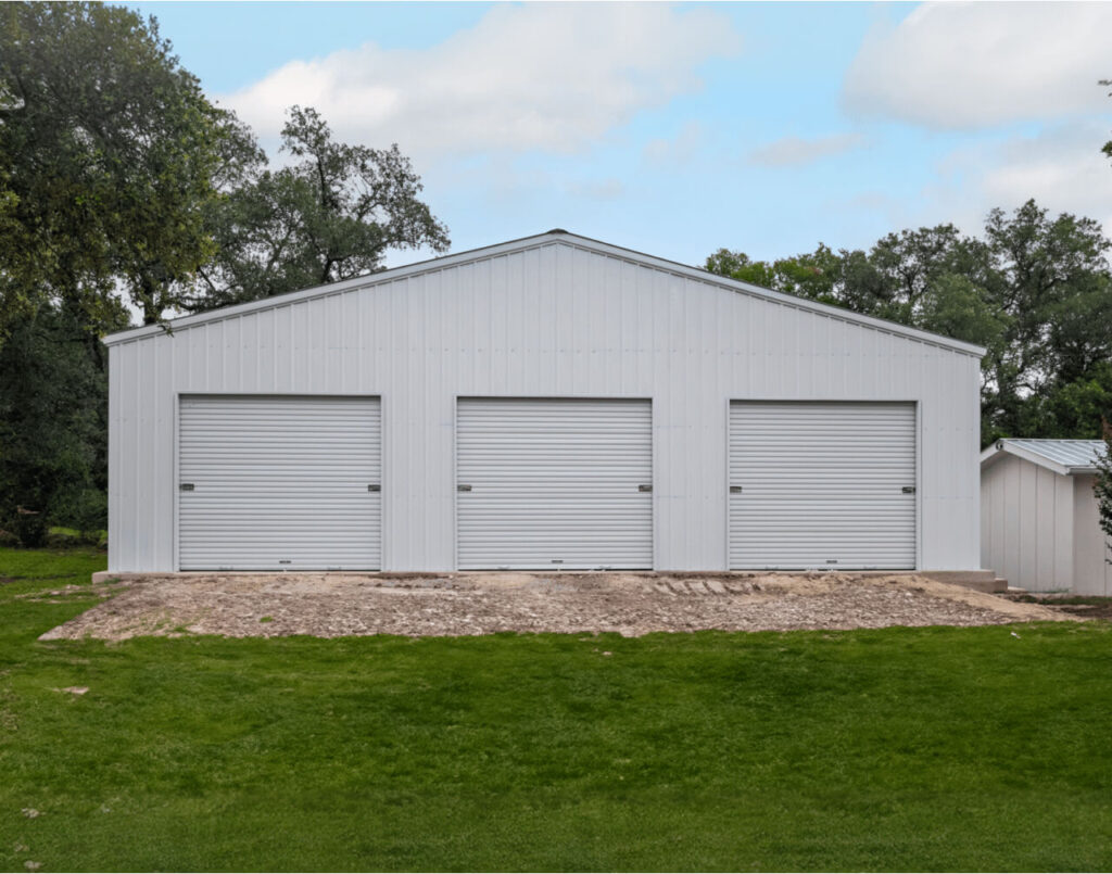 Large white metal warehouse building with three roll-up bay doors on a wide front facade, showing the kind of open-span commercial storage structure suited for vehicle storage, equipment access, or multi-bay warehouse operations.