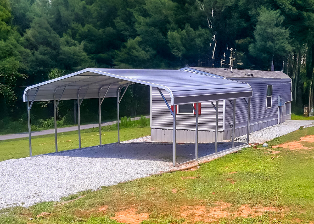 Metal RV carport attached to a mobile home with a gravel driveway and wooded background.