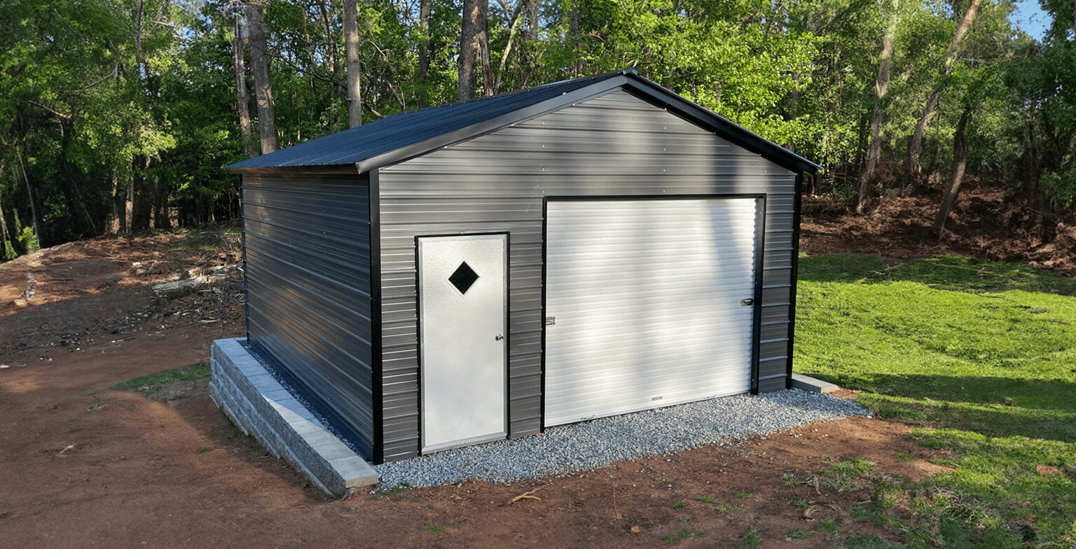 Small dark-gray metal barn with a gable roof set in a wooded area, combining a large roll-up door and side access door, showcasing a durable, low-maintenance structure suited for animal shelter, feed storage, or a compact workshop.