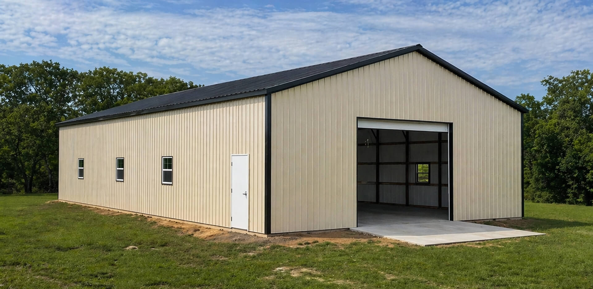Tan metal warehouse building with black trim and vertical roof on a rural property, featuring an open roll-up overhead door, walk-in personnel door, and side windows for natural light