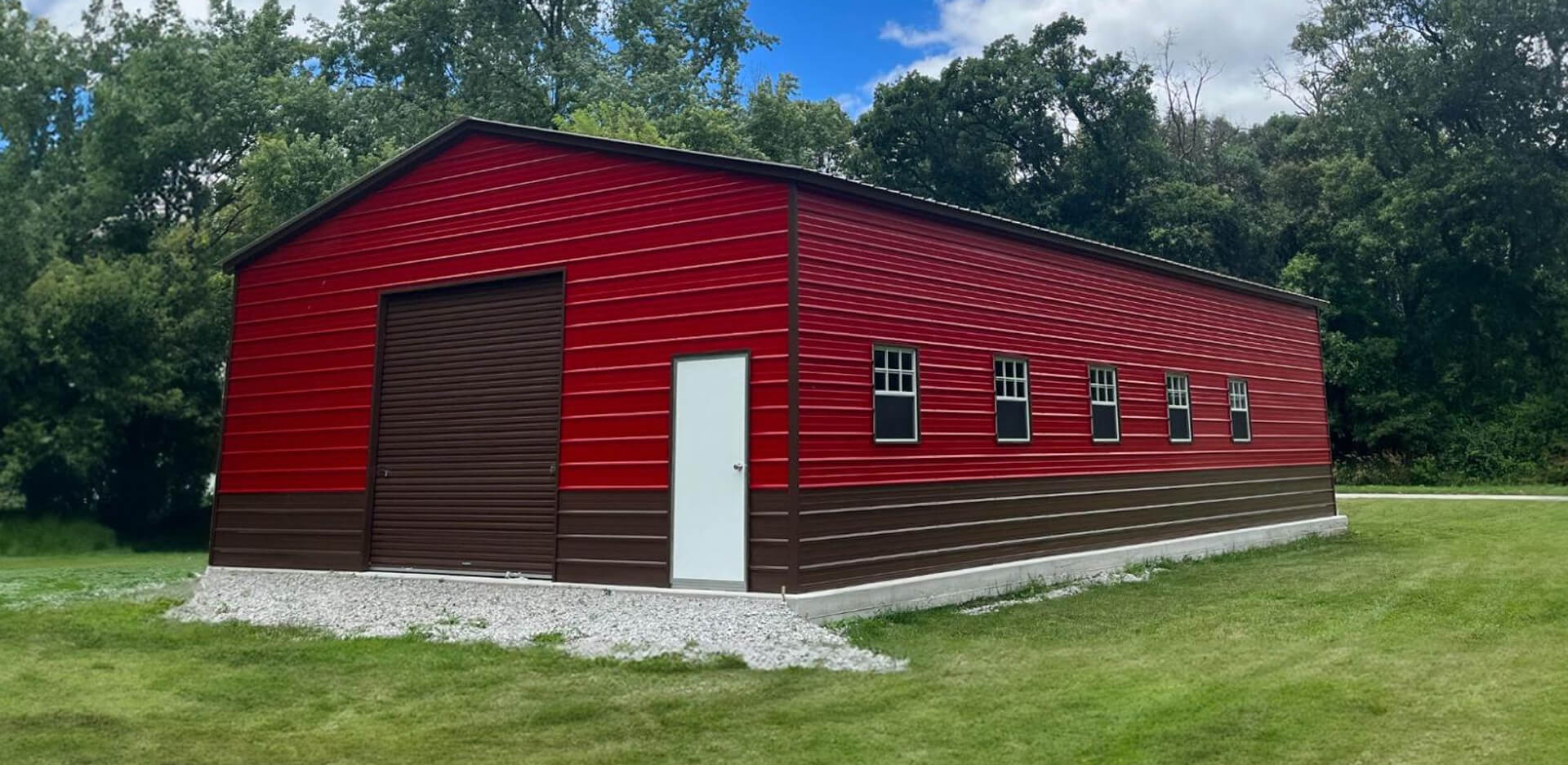 Large red and brown two-tone metal warehouse building with vertical roof, brown roll-up overhead door, white walk-in personnel door, and a row of five side windows, set on a gravel pad in a grassy clearing surrounded by trees