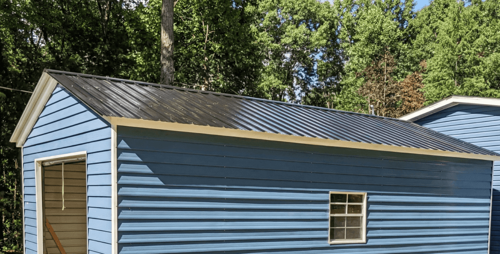 Blue metal storage shed with horizontal lap siding, black ribbed metal roof, white trim, and side window, set in a wooded backyard for durable outdoor storage.