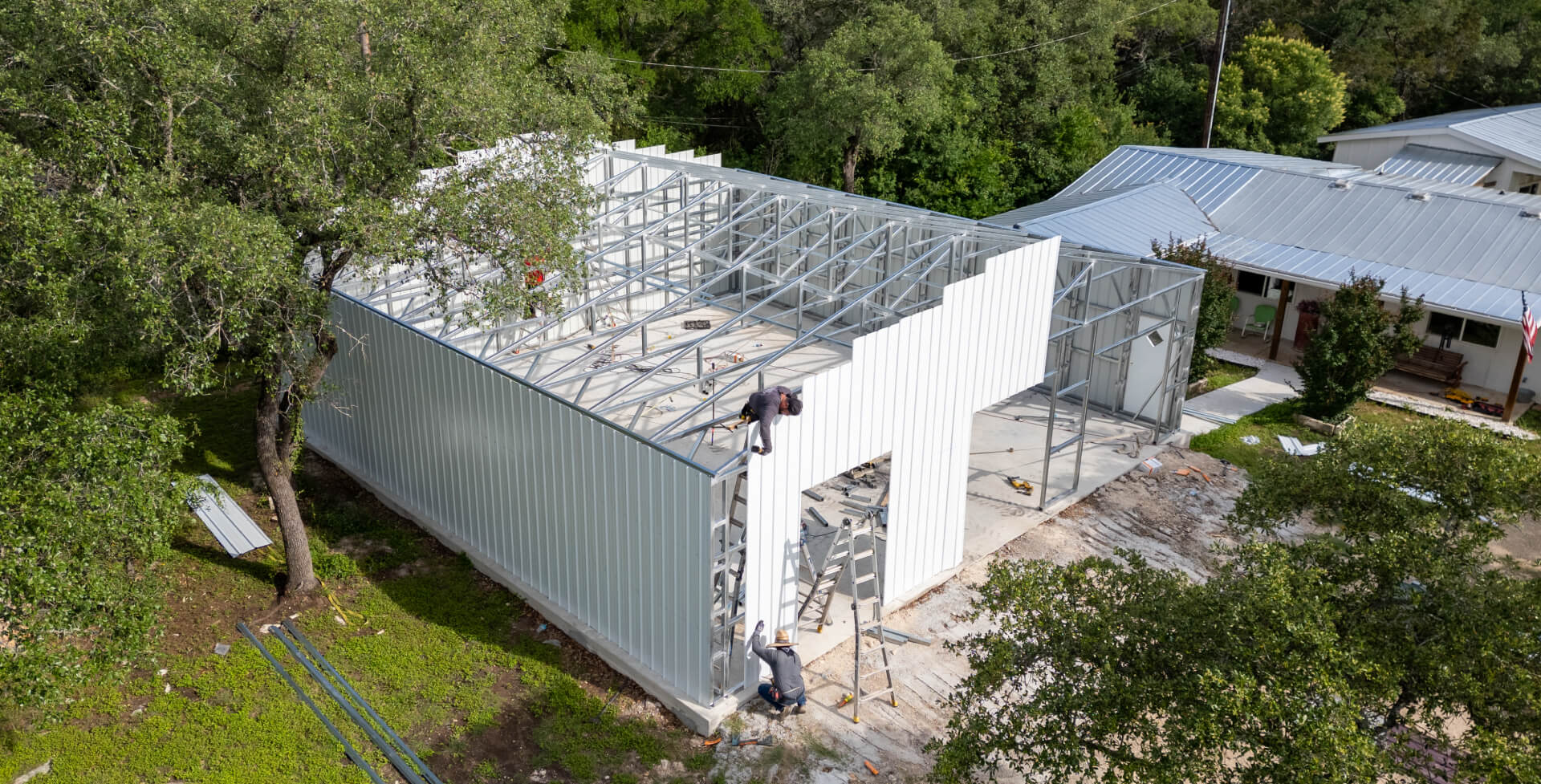 Large steel metal building under construction with exposed framing and vertical panels being installed, showing step-by-step prefab garage assembly process.