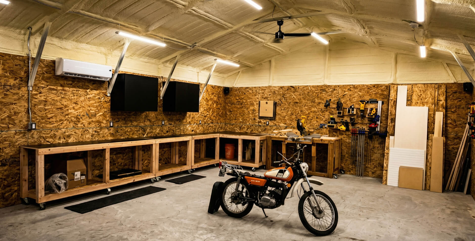 Insulated metal garage interior with spray foam ceiling, wood workbenches, mounted cabinets, power tools, and motorcycle, designed as a functional home workshop.