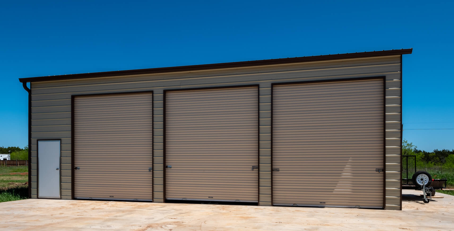 Tan steel garage building with three large roll-up doors and side entry door, ideal for equipment storage, vehicle parking, or backyard workshop space.
