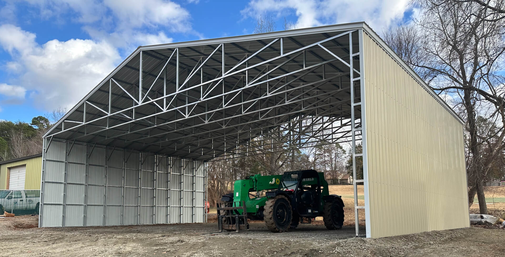 A large open-sided metal building with a peaked roof and exposed steel trusses stands on a dirt lot, partially enclosed with metal panels on one end. A green telehandler is parked inside beneath the structure, surrounded by trees and a partly cloudy sky.