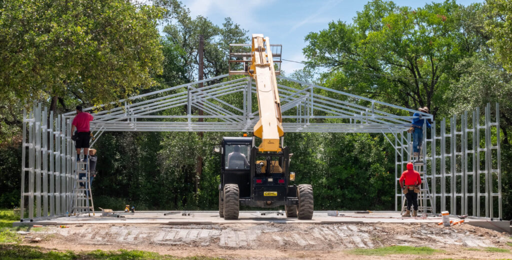 Construction crew assembling a steel building frame using a telescopic lift, showing truss placement during prefab metal garage installation.