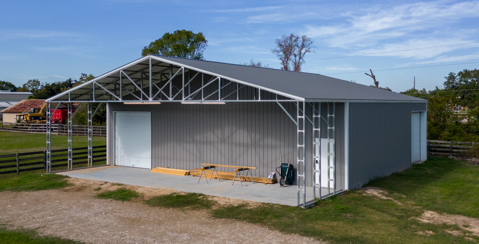 A gray metal building with an attached open lean-to features a roll-up garage door and exposed steel framing along the front corner. Lumber and tools rest on the concrete slab, with a fenced yard and rural landscape in the background.