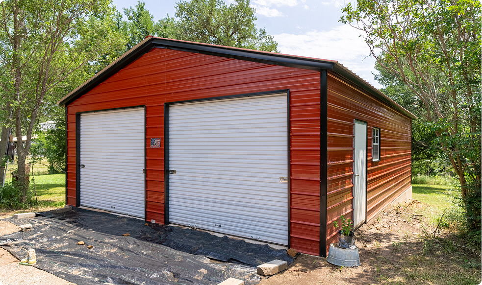 Two-car red metal garage with white roll-up doors and side entrance on a gravel lot