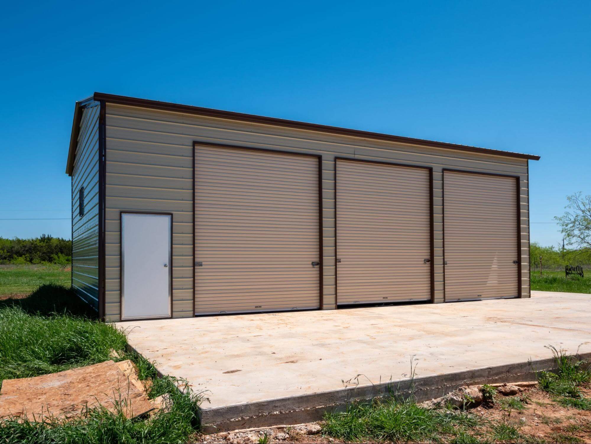 A 30×40 vertical-roof metal garage with three large roll-up doors and a side entry door, installed on a poured concrete slab in an open rural setting, illustrating a three-bay metal building configuration ideal for vehicle storage or a workshop.