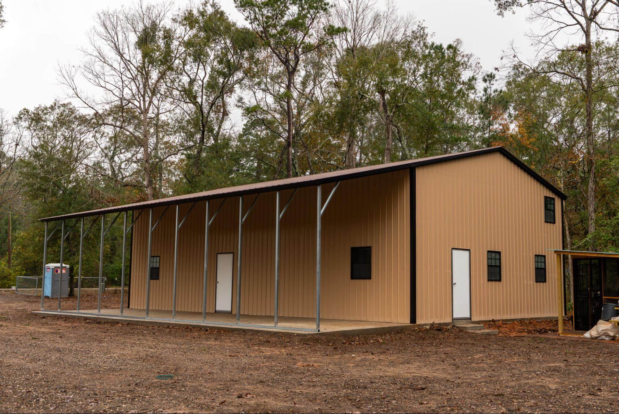 A large tan metal building with brown trim featuring an attached lean-to overhang supported by steel posts, demonstrating a versatile metal building configuration with covered workspace alongside enclosed storage.