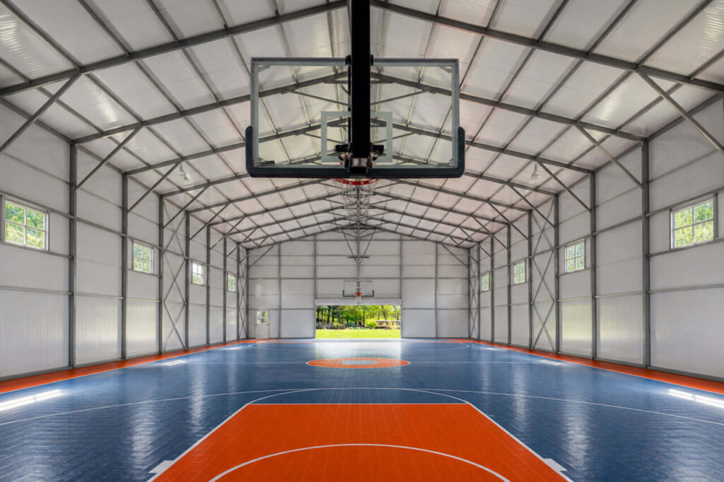 Interior view of a metal building converted into an indoor basketball court, featuring a vibrant blue and orange playing surface with white markings, a glass backboard and hoop in the foreground, and exposed gray steel trusses and framing along the high vaulted ceiling. Daylight streams in through small windows along the side walls and an open doorway at the far end.