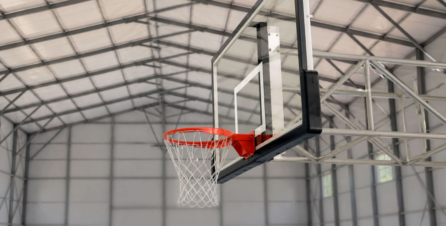 Close-up view of a basketball hoop with a clear glass backboard, orange rim, and white net mounted inside a metal building, with the gray steel roof trusses and framework visible in the background.