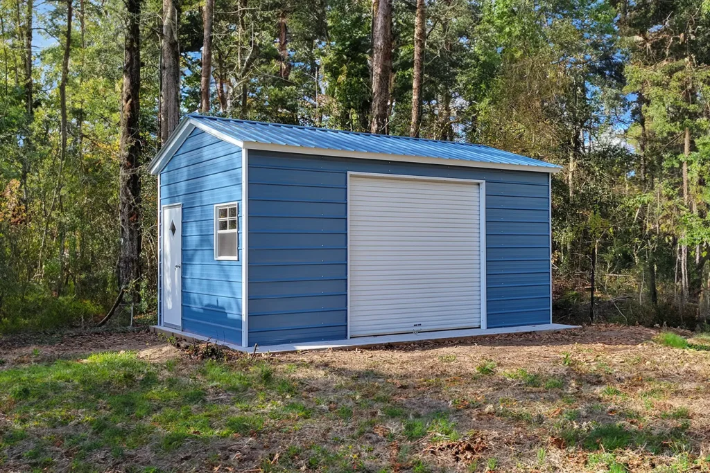 Small blue metal garage with white trim and a gabled roof, featuring a single white roll-up garage door, a white side entry door with a diamond window, and a small window. The structure sits on a dirt clearing surrounded by tall pine trees in a wooded area.