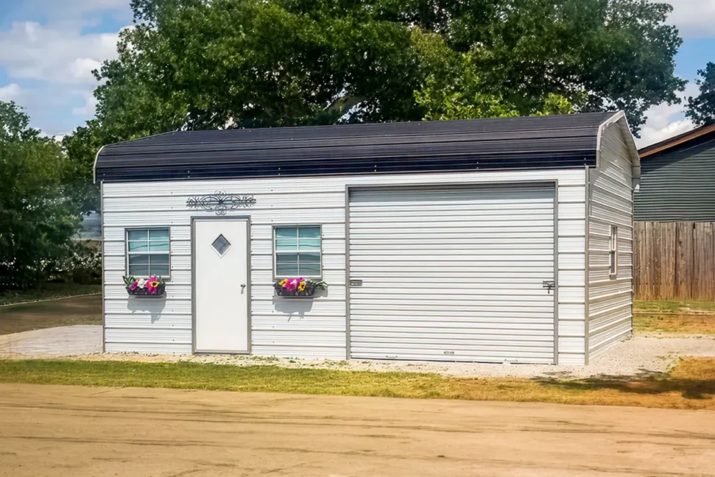 Small white metal garage with a curved black roof, featuring a white entry door with a diamond window, two windows with colorful flower boxes, a decorative scrollwork accent above the door, and a white roll-up garage door. The structure sits on a gravel pad with a wooden fence on one side and a large tree in the background.