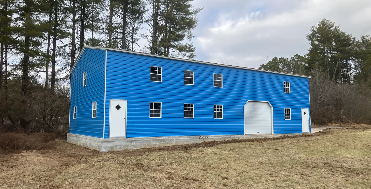 Large two-story bright blue metal barndominium with white trim, featuring multiple white-framed windows on both levels, two white entry doors with diamond windows, and a white roll-up garage door. The structure sits on a concrete foundation in a wooded clearing under a partly cloudy sky.