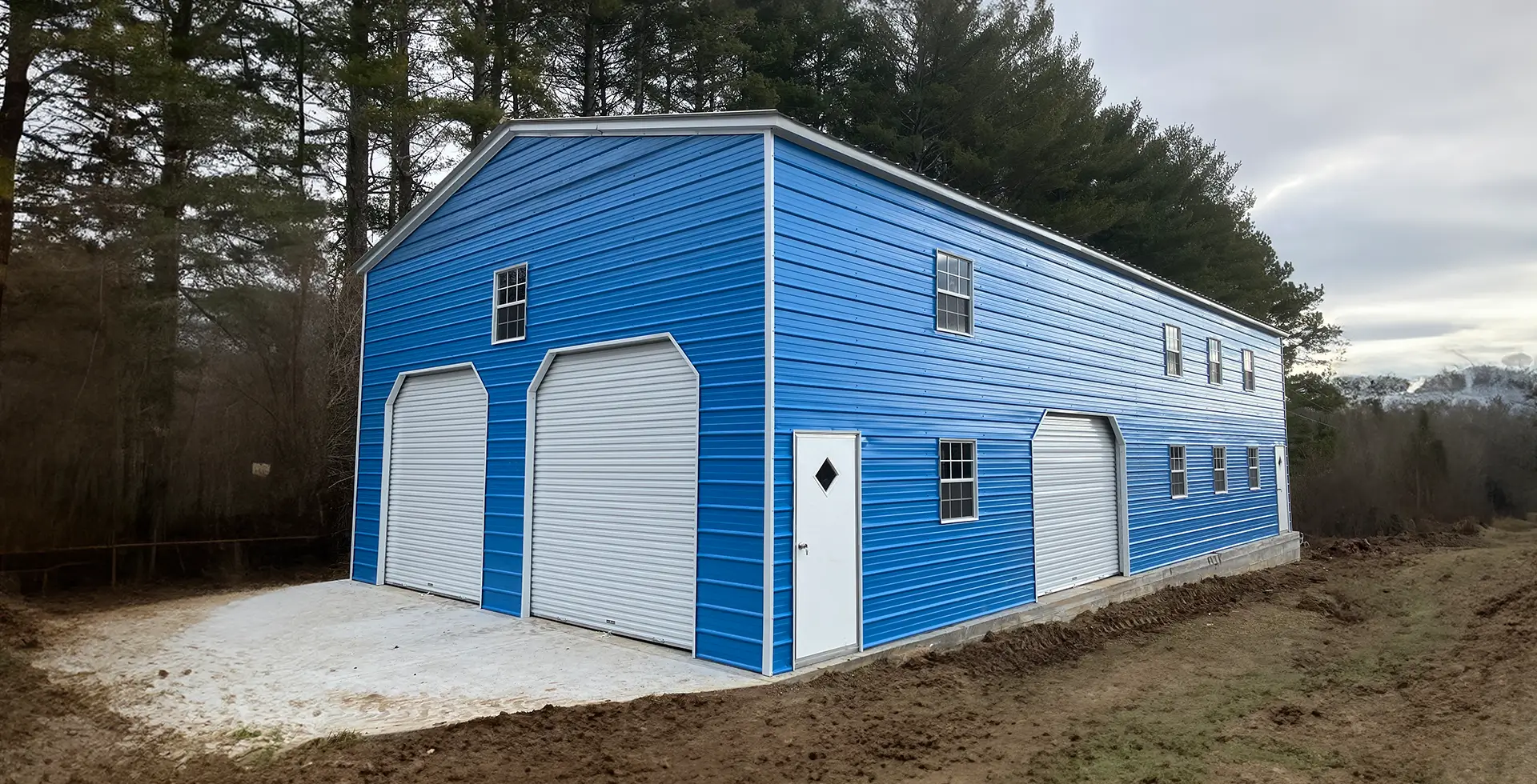 Large two-story bright blue metal barndominium with white trim and a gambrel-style roof, featuring two white roll-up garage doors on the front, additional roll-up doors and entry doors along the side, multiple white-framed windows on both levels, and a white side entry door with a diamond window. The structure sits on a concrete foundation in a wooded area with bare trees and disturbed earth around it.