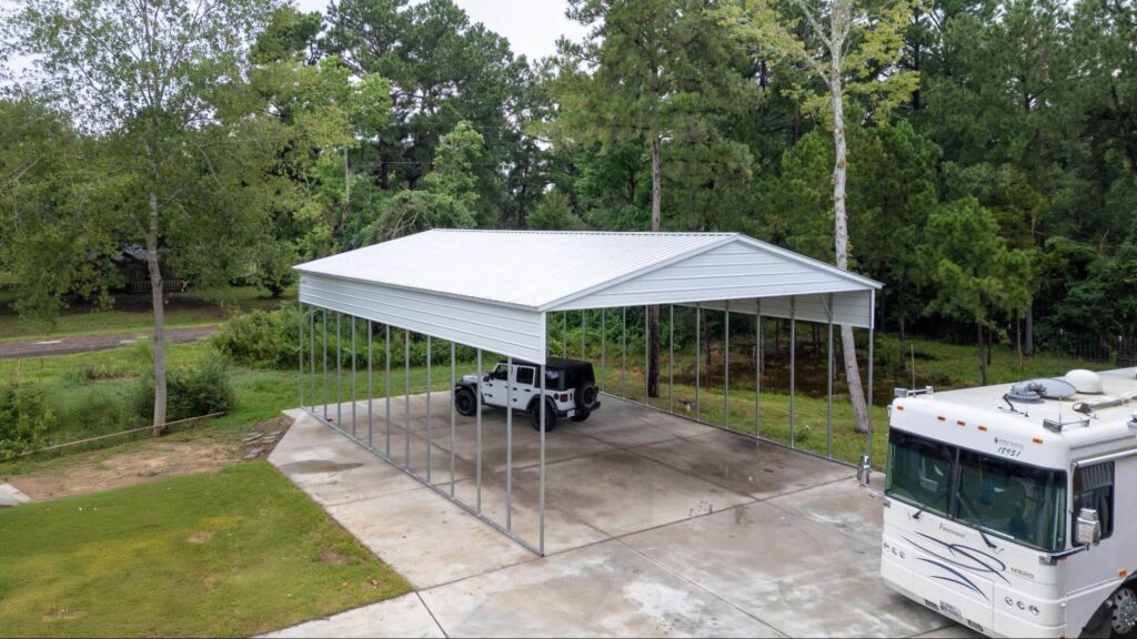 A large white metal carport with a peaked roof installed over a finished concrete slab, sheltering a Jeep Wrangler with an RV parked alongside, demonstrating proper slab dimensions and anchoring for a vehicle storage building.