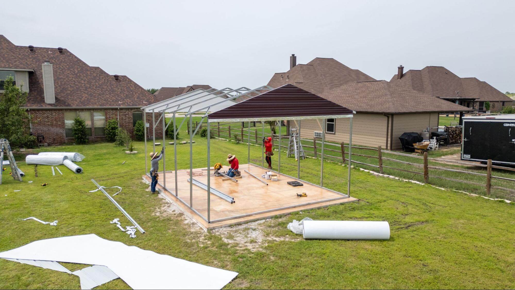A professional crew assembles a metal carport frame on a finished concrete slab in a suburban backyard, illustrating the value of hiring a concrete contractor for larger slab projects that anchor a metal building.