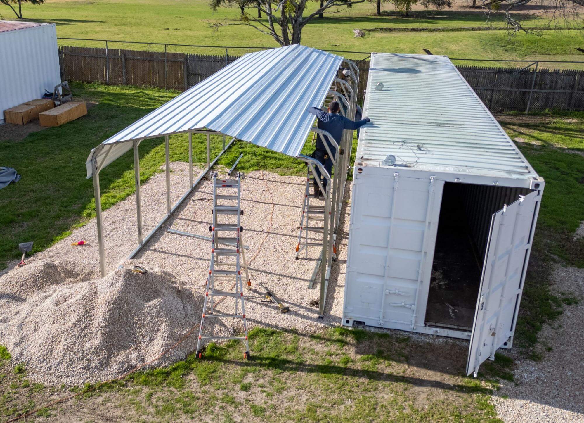 A metal carport being installed over a gravel base alongside a shipping container, showing an alternative foundation option to a concrete slab for lighter-duty metal building applications.