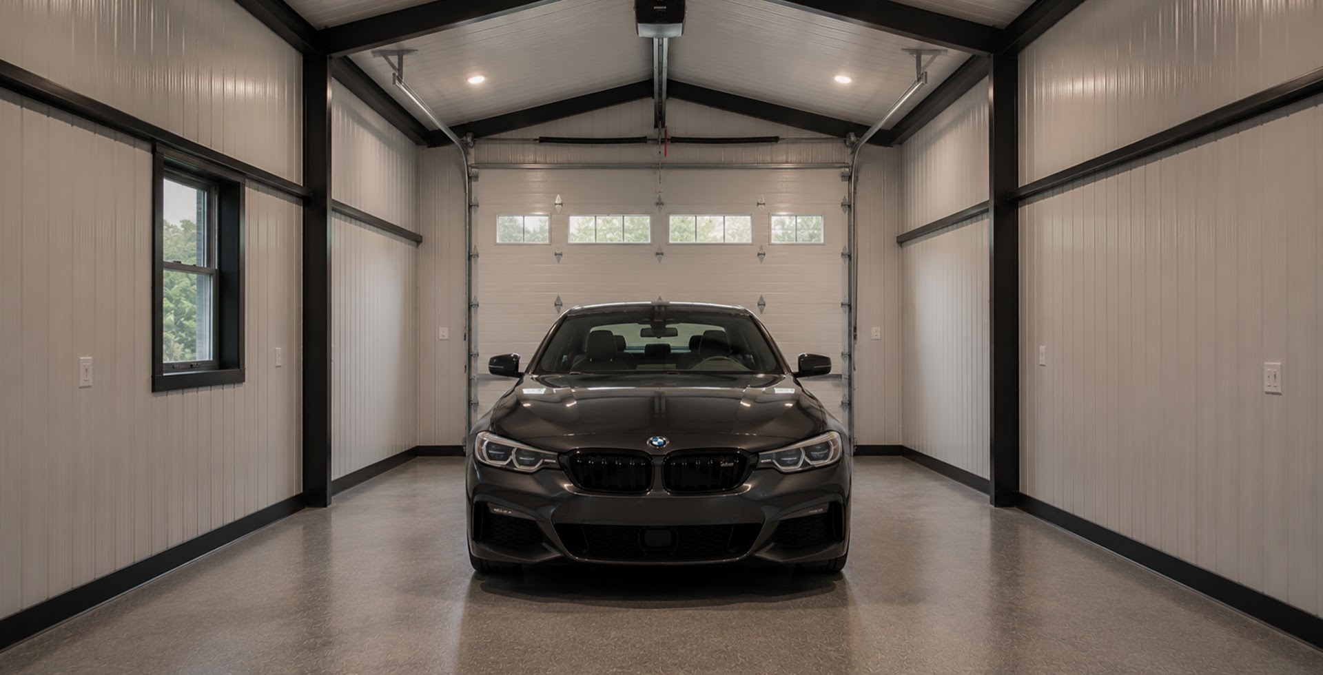 Interior of a residential metal garage with white walls, exposed black steel trusses, polished concrete floors, and a row of small transom windows along the back wall above the garage door. A dark gray BMW sedan is parked in the center of the bay.