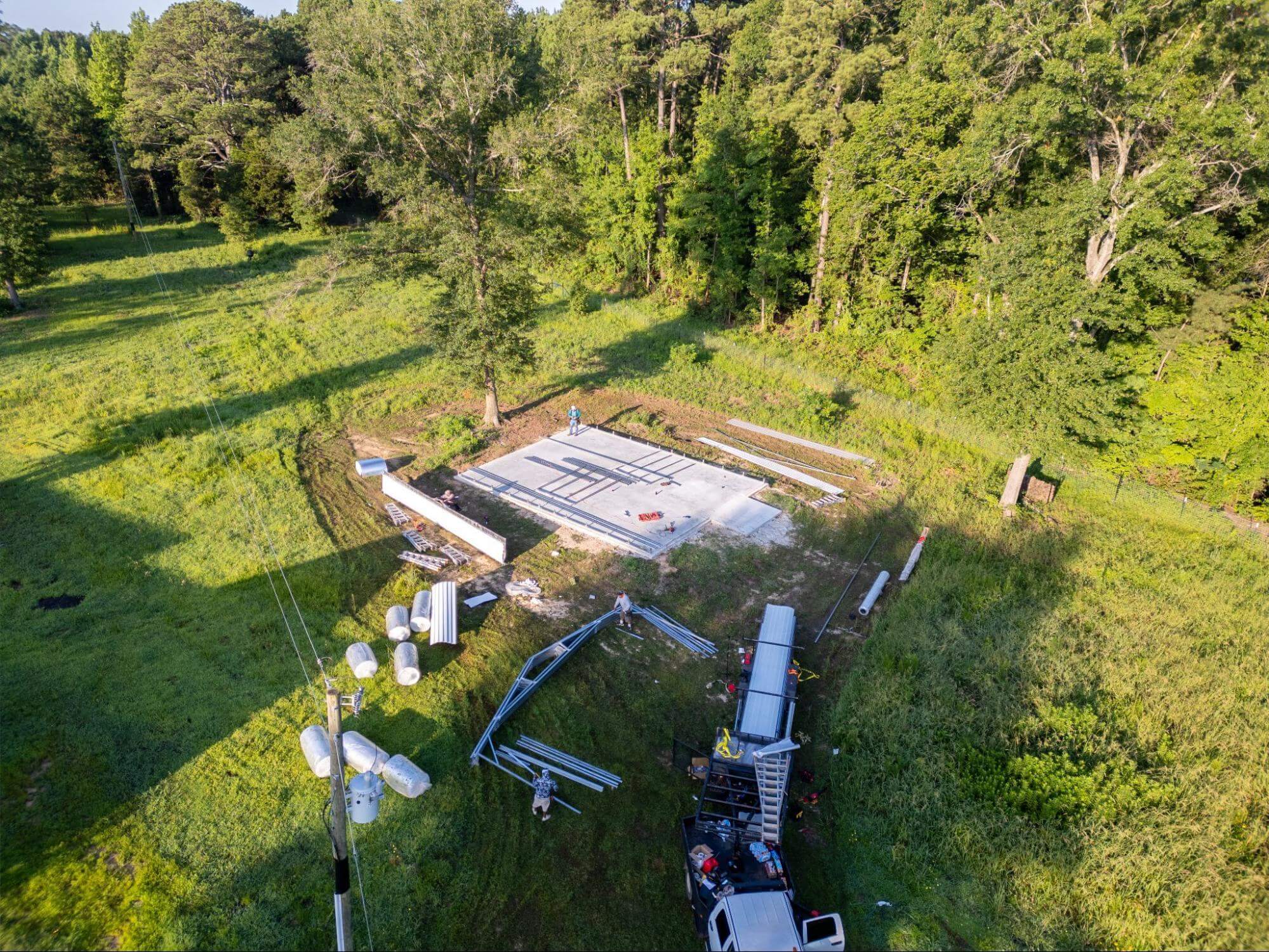 Aerial view of a freshly poured concrete slab foundation in a wooded clearing, with metal building framing components and an installation crew preparing to assemble a metal building on top.